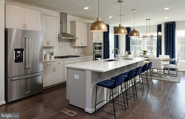 kitchen with a kitchen island with sink, stainless steel appliances, a sink, hanging light fixtures, and light countertops