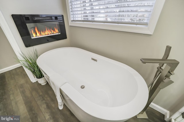 bathroom featuring a glass covered fireplace, a freestanding bath, baseboards, and wood finished floors