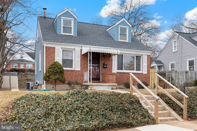 cape cod home with central air condition unit, a shingled roof, fence, and brick siding