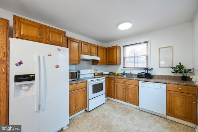 kitchen with brown cabinets, dark countertops, a sink, white appliances, and under cabinet range hood