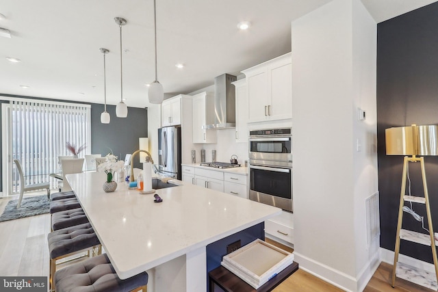 kitchen with stainless steel appliances, a sink, a kitchen breakfast bar, wall chimney exhaust hood, and light wood finished floors