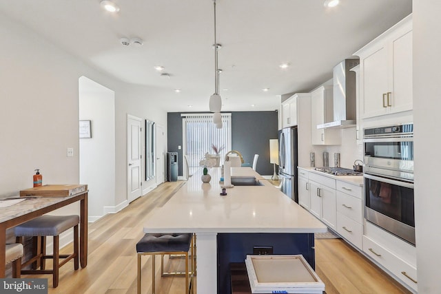 kitchen with stainless steel appliances, a sink, white cabinetry, wall chimney range hood, and an island with sink