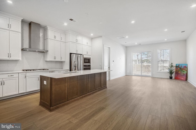 kitchen featuring a center island with sink, light countertops, decorative backsplash, appliances with stainless steel finishes, and wall chimney exhaust hood