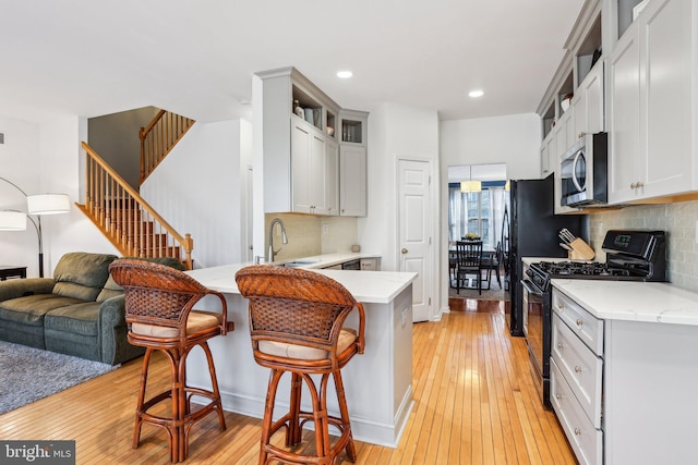 kitchen with black range with gas cooktop, a peninsula, a sink, light wood finished floors, and stainless steel microwave