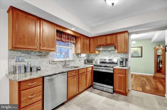 kitchen featuring under cabinet range hood, a sink, appliances with stainless steel finishes, light stone countertops, and brown cabinetry