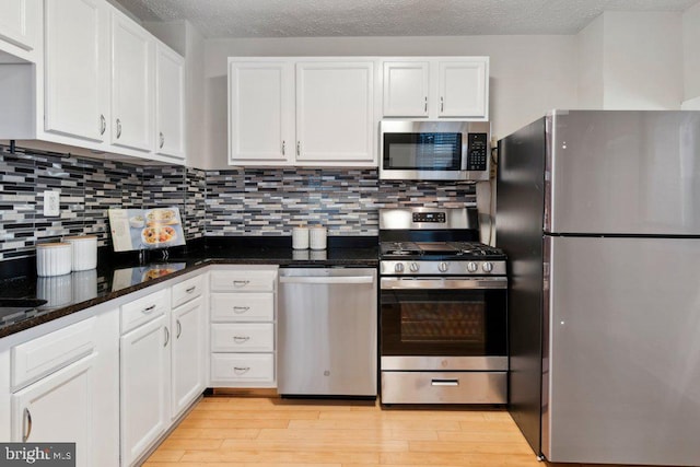 kitchen with backsplash, appliances with stainless steel finishes, white cabinetry, dark stone countertops, and light wood-type flooring