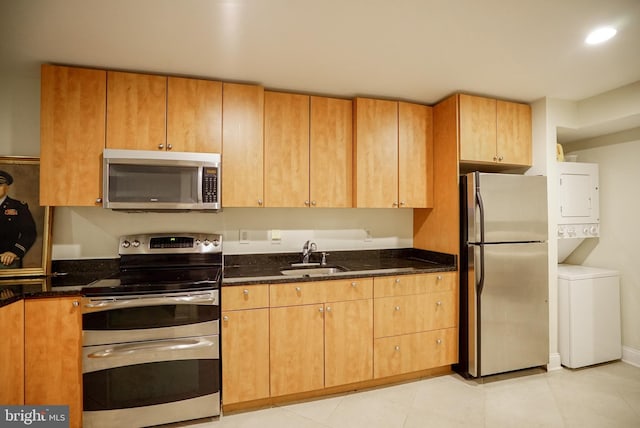 kitchen with a sink, dark stone counters, appliances with stainless steel finishes, stacked washer / dryer, and light tile patterned floors