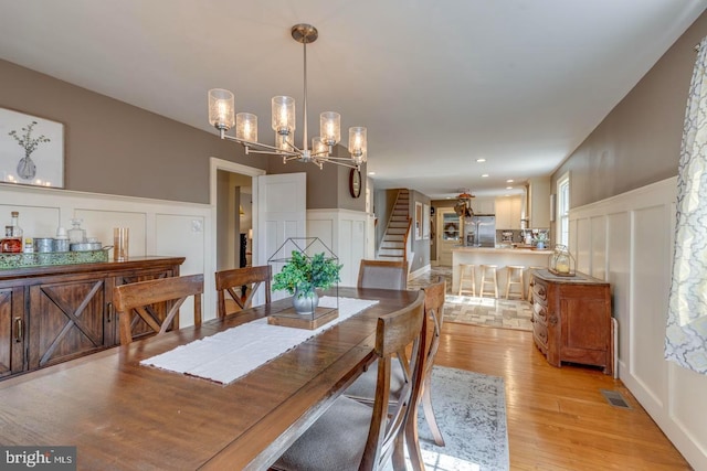 dining room with a wainscoted wall, stairs, light wood-style floors, a chandelier, and a decorative wall