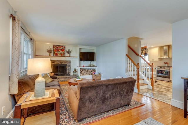 living area featuring stairs, a stone fireplace, light wood-type flooring, and baseboards