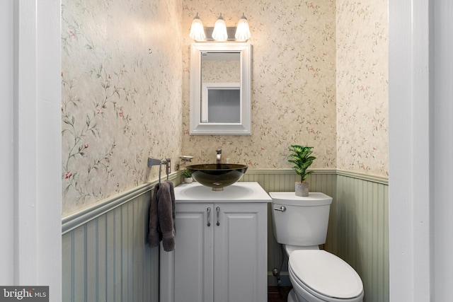 half bath with a wainscoted wall, vanity, toilet, and wallpapered walls