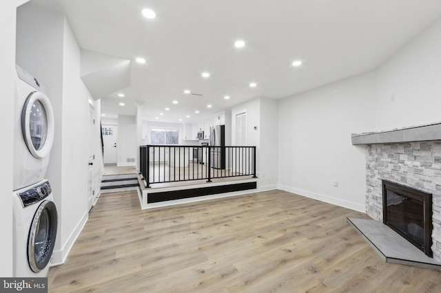 clothes washing area featuring light wood finished floors, stacked washer / drying machine, recessed lighting, a stone fireplace, and baseboards