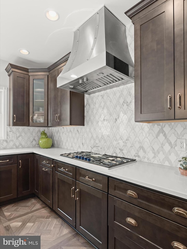 kitchen featuring dark brown cabinets, glass insert cabinets, wall chimney range hood, light countertops, and stainless steel gas stovetop