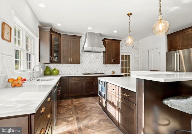 kitchen featuring wall chimney range hood, dark brown cabinetry, appliances with stainless steel finishes, a sink, and light countertops
