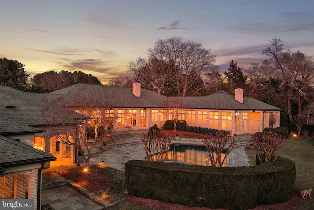 back of house at dusk with a chimney, an outdoor pool, and a patio