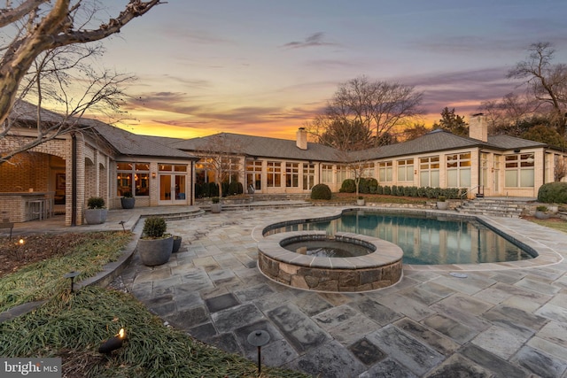 back of property at dusk featuring a patio area, a pool with connected hot tub, and a chimney