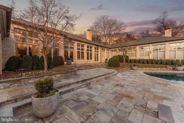 back of property at dusk featuring an outdoor pool, a patio area, brick siding, and a chimney