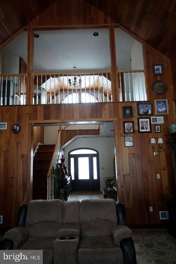 carpeted living area with high vaulted ceiling, stairs, wooden walls, and visible vents