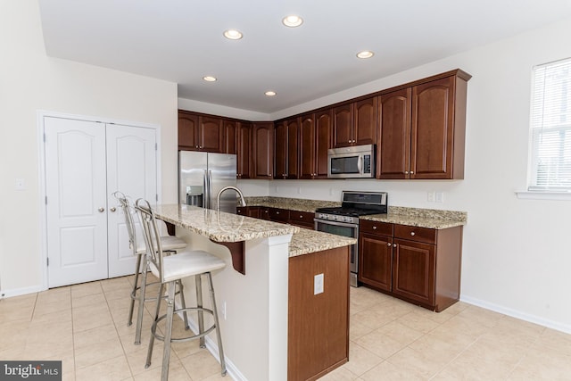 kitchen with light stone counters, appliances with stainless steel finishes, dark brown cabinetry, an island with sink, and a kitchen bar