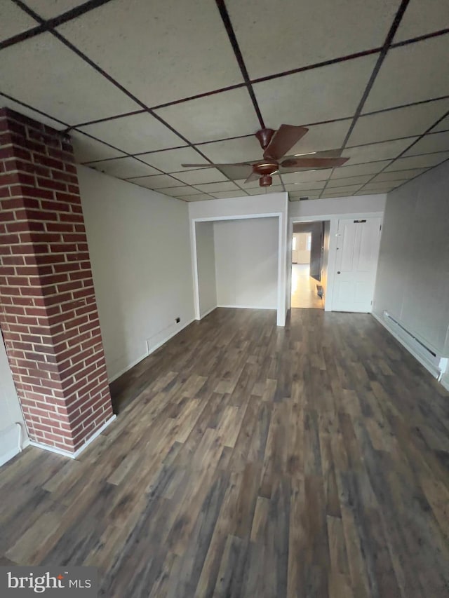 unfurnished living room with a baseboard radiator, dark wood-type flooring, a ceiling fan, brick wall, and a drop ceiling