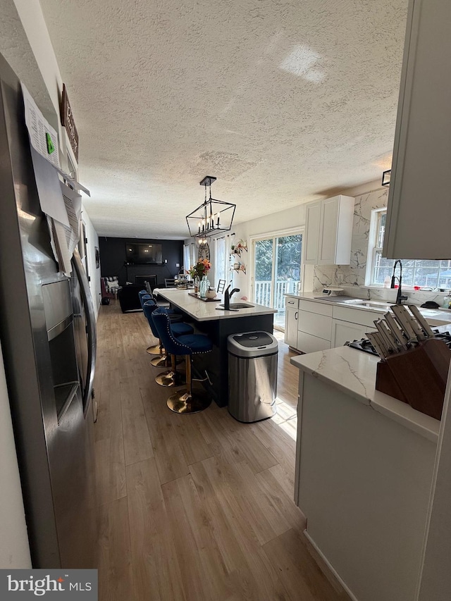 kitchen with pendant lighting, stainless steel refrigerator with ice dispenser, light wood-style flooring, white cabinetry, and an island with sink