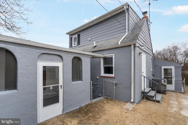 rear view of property featuring a shingled roof, central air condition unit, a chimney, and stucco siding