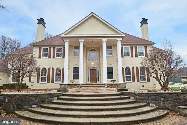 greek revival inspired property with a chimney, roof with shingles, and stucco siding