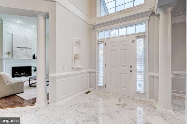 foyer featuring a decorative wall, a towering ceiling, marble finish floor, a brick fireplace, and ornate columns