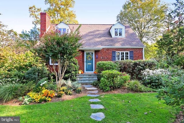 cape cod home featuring a shingled roof, a front yard, a chimney, and brick siding