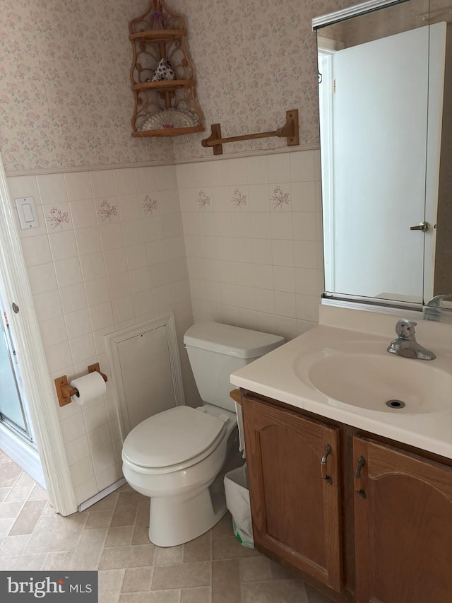 bathroom with wallpapered walls, wainscoting, toilet, vanity, and tile walls