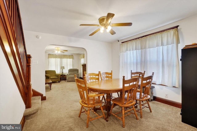 dining space featuring arched walkways, light colored carpet, stairway, ceiling fan, and baseboards