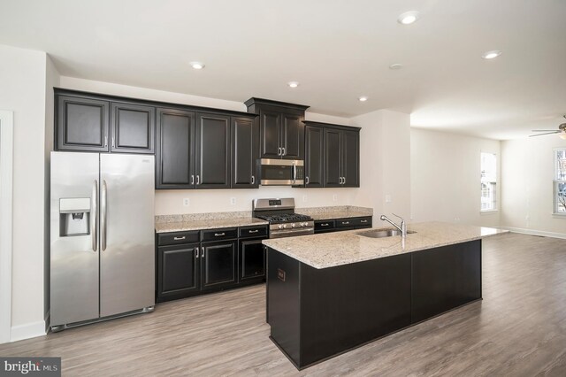 kitchen featuring light wood-style flooring, appliances with stainless steel finishes, a kitchen island with sink, dark cabinetry, and a sink