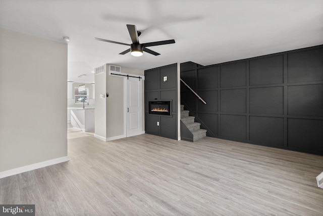 unfurnished living room with a barn door, a ceiling fan, stairway, light wood-style floors, and a decorative wall