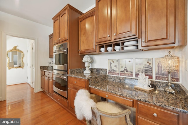 kitchen featuring brown cabinetry, stainless steel double oven, light wood-type flooring, and dark stone countertops