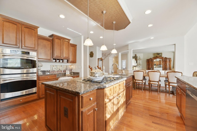 kitchen with stainless steel appliances, hanging light fixtures, brown cabinets, dark stone counters, and an island with sink