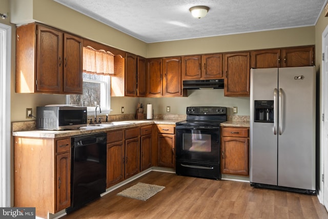 kitchen with under cabinet range hood, a sink, light countertops, black appliances, and light wood finished floors