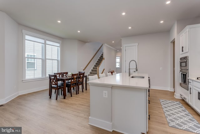 kitchen with an island with sink, light wood-style flooring, white cabinetry, a sink, and stainless steel double oven
