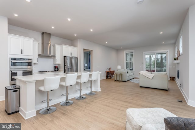 kitchen featuring wall chimney exhaust hood, open floor plan, light wood-type flooring, a spacious island, and stainless steel appliances