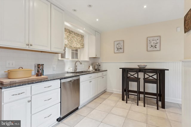 kitchen with light tile patterned floors, white cabinets, stainless steel dishwasher, wainscoting, and dark stone counters