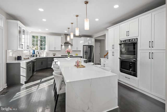 kitchen featuring a center island, wall chimney range hood, dishwasher, double wall oven, and stainless steel fridge