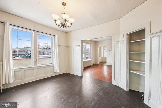 unfurnished dining area with arched walkways, a notable chandelier, radiator, a decorative wall, and dark wood-type flooring