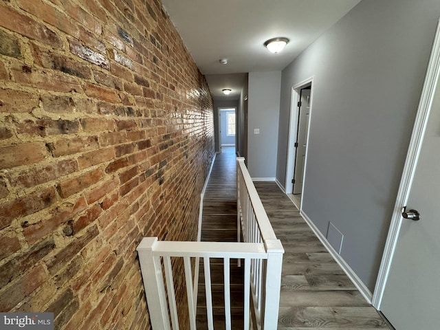 hallway with brick wall, dark wood-style flooring, visible vents, an upstairs landing, and baseboards