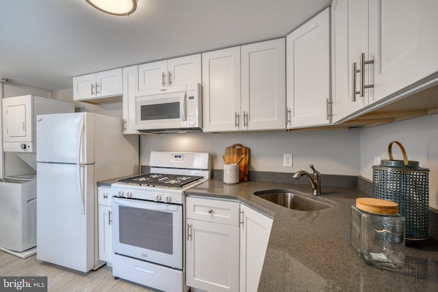 kitchen featuring stacked washer / dryer, white cabinets, a sink, dark stone counters, and white appliances