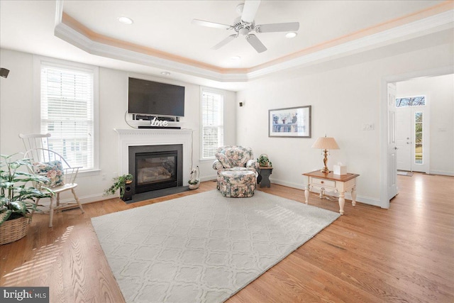 living area featuring a tray ceiling, light wood-style flooring, a fireplace with flush hearth, ceiling fan, and baseboards