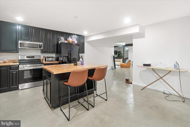 kitchen featuring stainless steel appliances, concrete floors, wood counters, dark cabinetry, and a kitchen bar