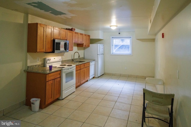 kitchen with brown cabinetry, light tile patterned flooring, a sink, dark stone counters, and white appliances