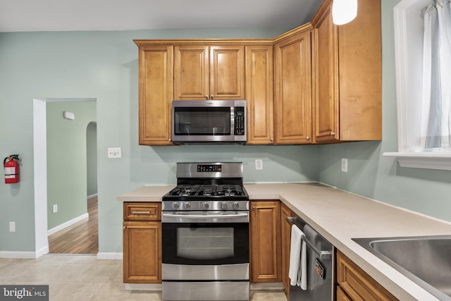 kitchen featuring arched walkways, stainless steel appliances, baseboards, light countertops, and brown cabinetry