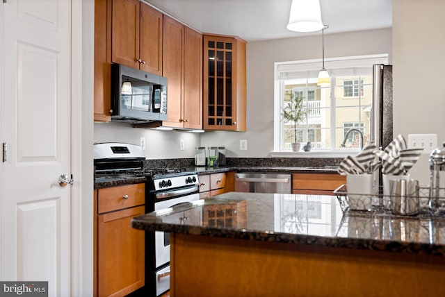 kitchen featuring decorative light fixtures, stainless steel appliances, brown cabinetry, glass insert cabinets, and dark stone countertops