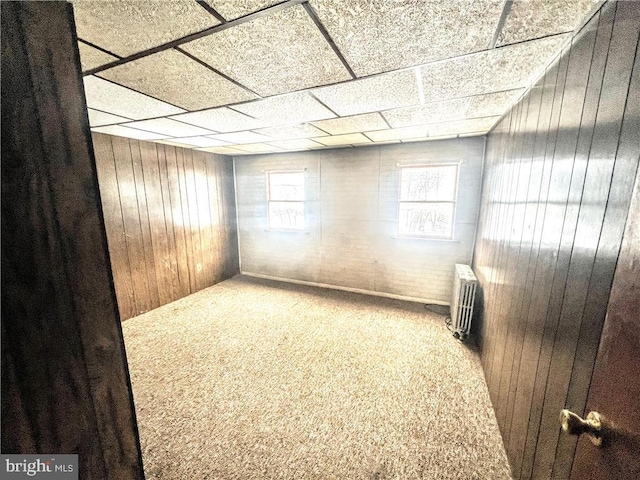 unfurnished room featuring carpet floors, a drop ceiling, radiator, and wooden walls