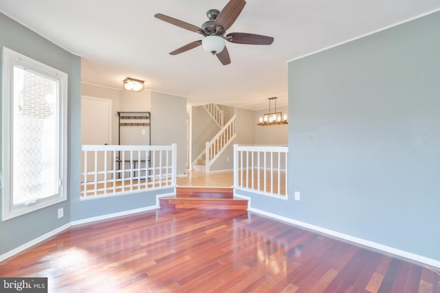 empty room with ceiling fan with notable chandelier, wood finished floors, baseboards, and stairs