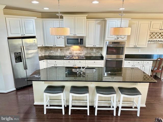 kitchen featuring a kitchen island with sink, a sink, appliances with stainless steel finishes, dark stone countertops, and pendant lighting